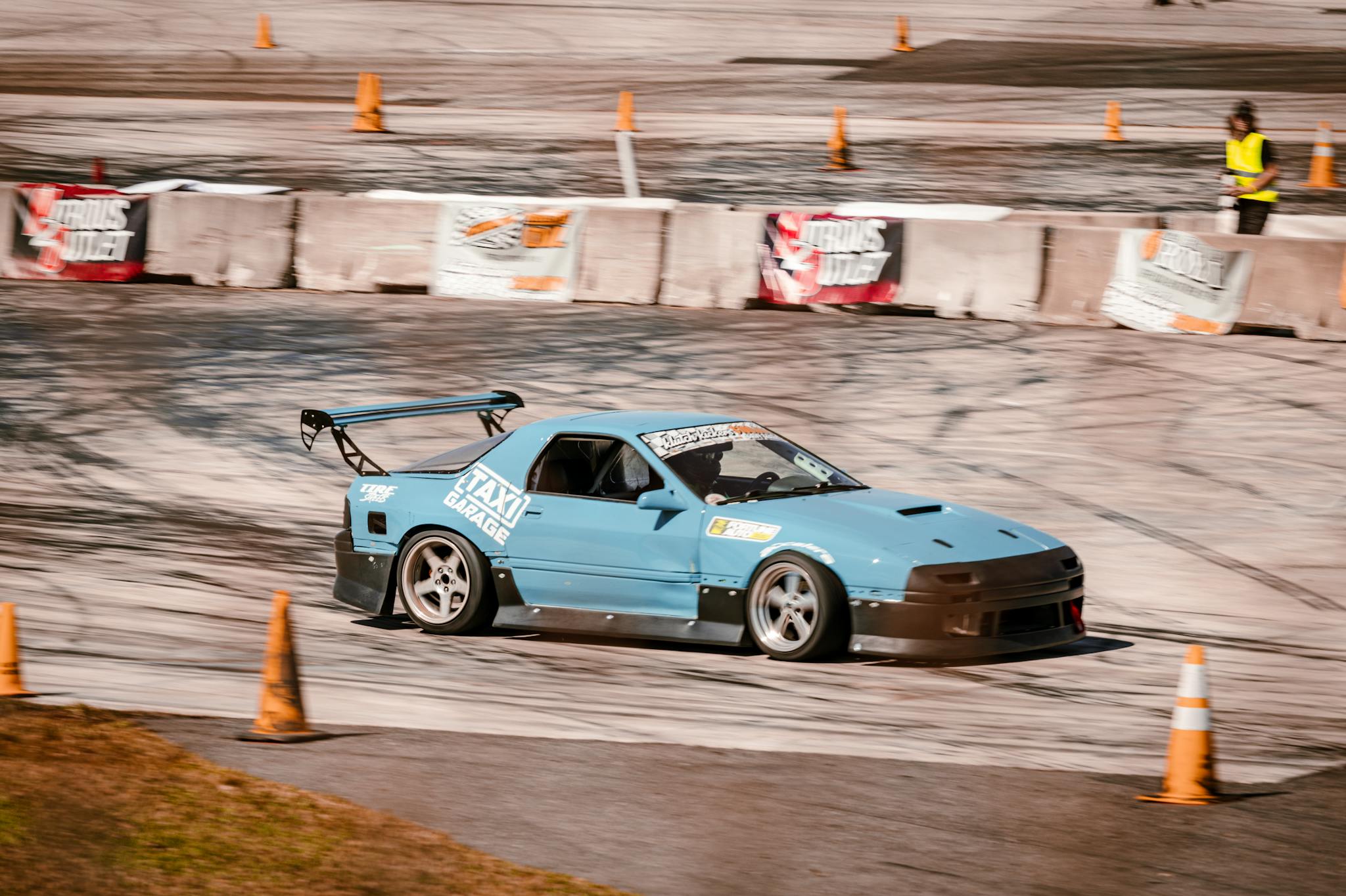 A blue sports car performing a drift maneuver on a race track with spectators nearby.