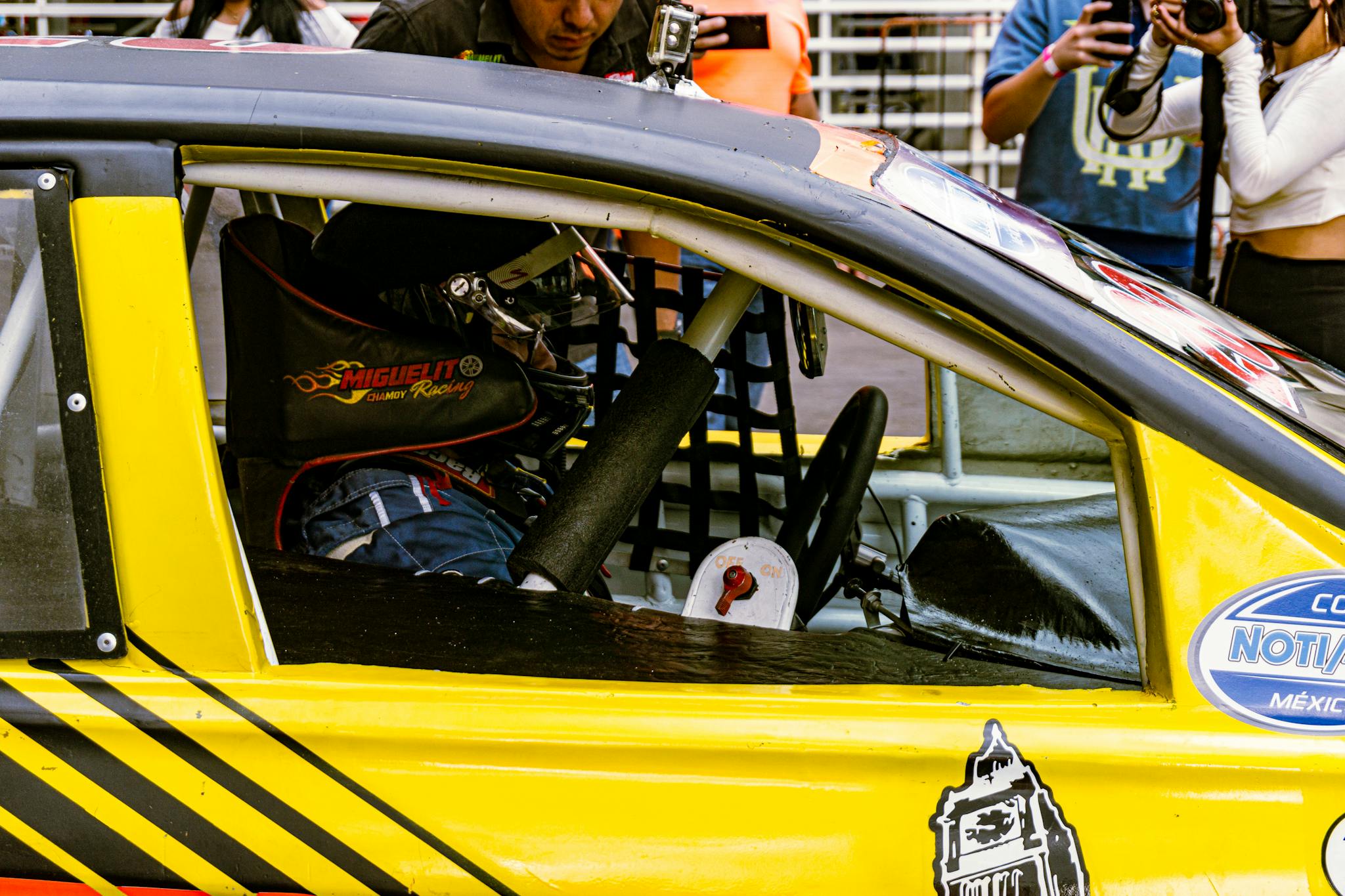 A professional driver wearing a helmet sits in a yellow race car during a motorsport event.